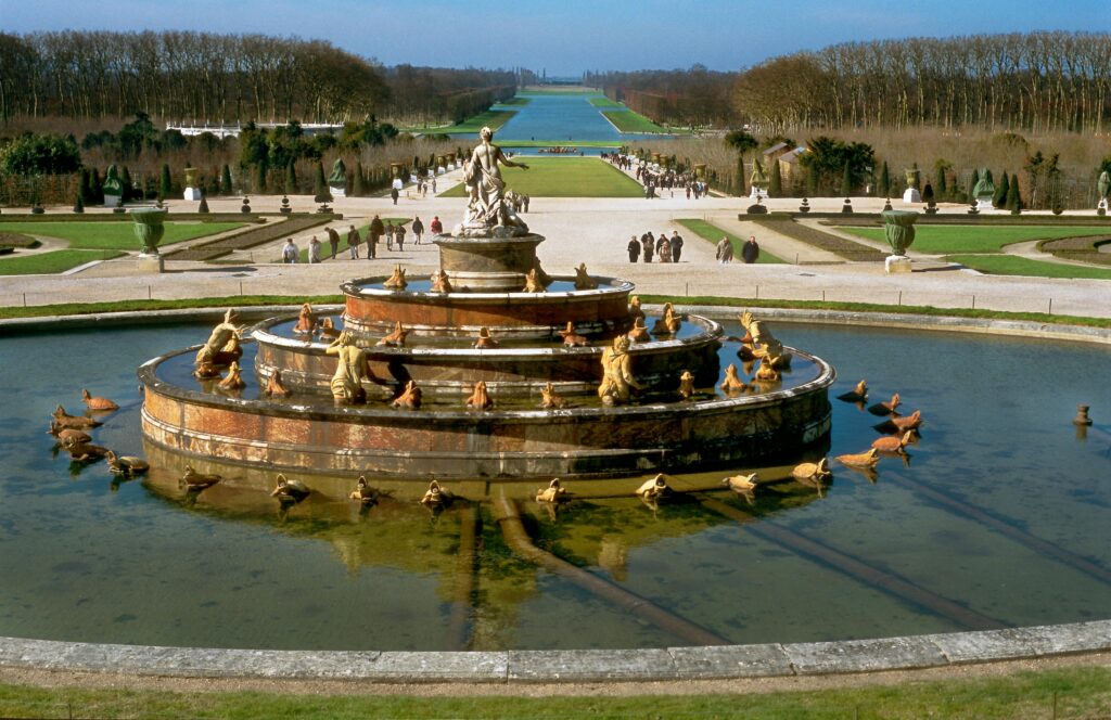 Classic fountain in the Gardens of Versailles surrounded by lush greenery and pathways.